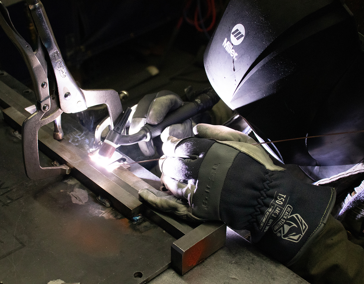 Welder using a Miller helmet and TIG torch to fuse a steel frame held in vise grips, sparks and white-hot arc light reflecting off the gloves.