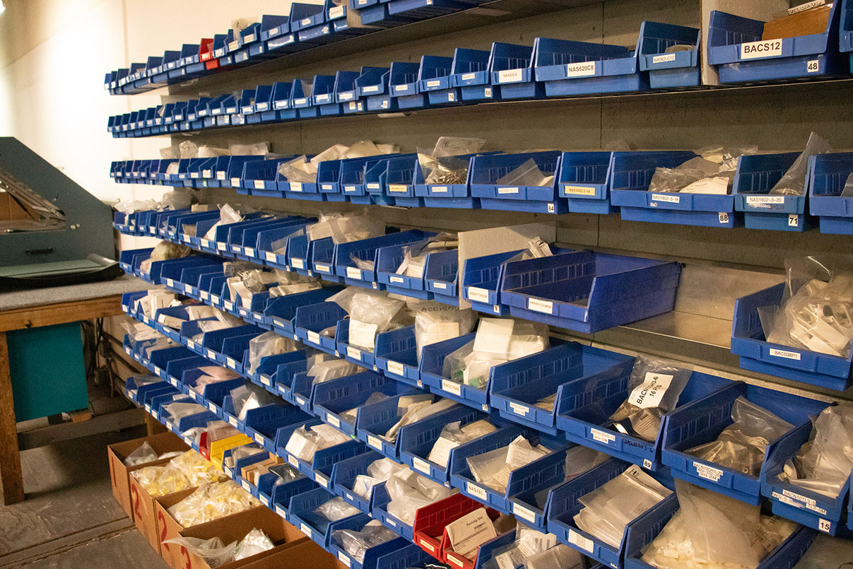 Aerospace hardware bins labeled with NAS, BACS, and BACN part numbers, stocked floor-to-ceiling along an inventory wall.