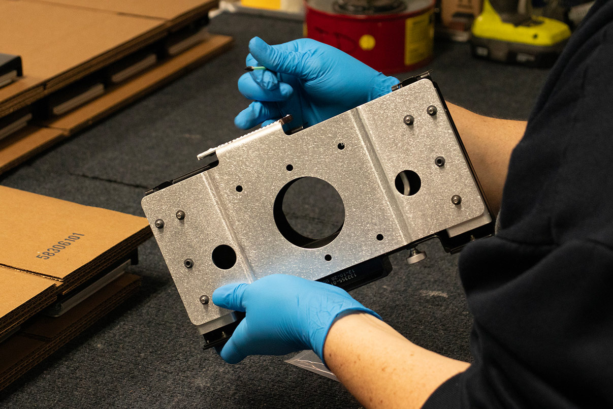 Technician in blue nitrile gloves holding a powder-coated aluminum mounting plate during final inspection.