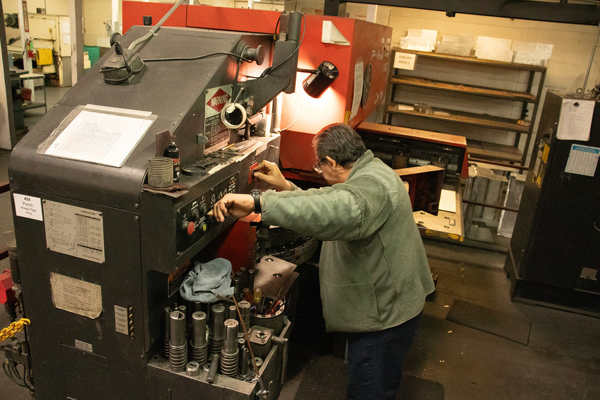 Operator running an Amada Pega turret punch press, positioned at the control panel with the punch tooling visible below.