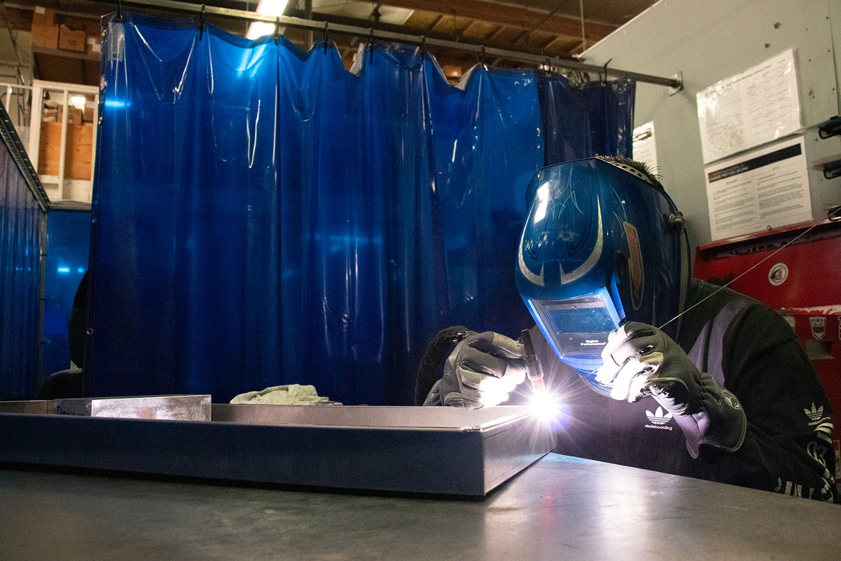 Welder behind a blue welding curtain, MIG welding a steel weldment under a custom flame-painted helmet.