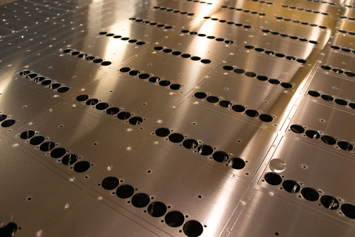 Tight overhead shot of a laser-cut sheet metal panel covered in perforated hole patterns, raking light catching the brushed aluminum surface.